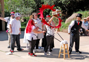 Photo by Rhonda Van Pelt. Professor Possible (Garfield Gerholdt) looks askance at the dragon his fellow performers have created. The mops are the creature’s front legs and its head was built from a cardboard box.